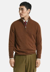Brauner Wollmischpullover mit Reißverschlusskragen, gerippten Bündchen und Saum. Über einem hellen karierten Hemd und einer dunklen Krawatte getragen, kombiniert mit grünen Hosen.
