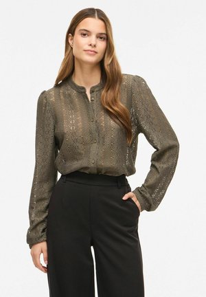 Jeune femme aux longs cheveux portant un chemisier en dentelle vert olive et un pantalon noir taille haute, debout avec une main dans la poche.