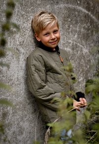 Giacca imbottita verde oliva con colletto nero, dotata di chiusura con zip e tiralampo in pelle marrone, indossata da un bambino davanti a un muro grigio.