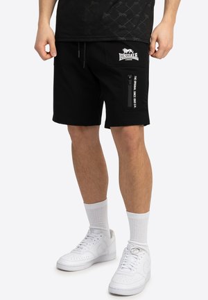 Čierne šortky vyrobené z mäkkej tkaniny, s vreckom na zips, bielym logom vpredu a elastickým pásom na šnúrku. Nosené s bielymi ponožkami a topánkami.