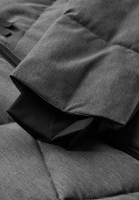 Veste grise isolée avec un design matelassé. La manche a un poignet intérieur noir, avec une finition lisse et texturée. Détails de fermeture éclair visibles.