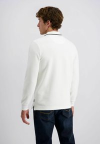 Man in een wit langamige polo-shirt met zwarte kraag en donkerblauwe jeans, met de rug naar de camera tegen een effen lichte achtergrond.