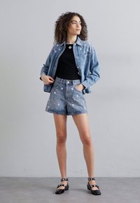Completo in denim di giacca e pantaloni corti di colore azzurro chiaro, con motivi floreali. Top nero sotto, abbinato a scarpe con strap nere. Texture liscia.