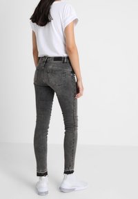Mujer con jeans ajustados negros desgastados, camiseta blanca de mangas enrolladas y zapatillas blancas, de pie frente a un fondo blanco.