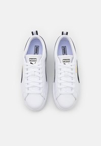 Baskets blanches avec des accents noirs, texture lisse, devant lacé et un label de logo sur la languette. Comprend un tirant au niveau du talon.