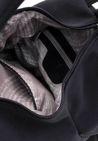 Interiör av svart perforerad tygpåse med ljusrosa foder och tryckt text. Inkluderar fack med dragkedja och mjuka avdelare.