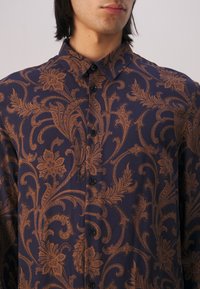 Homme portant une chemise bleu marine à boutons avec un motif floral et de vigne marron complexe, montré des épaules jusqu'au milieu de la poitrine.