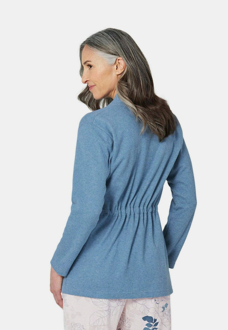 Helle blaue, langärmlige Jacke mit gerafftem Bund. Aus weichem Fleece-Material und mit einfachem Kragen-Design; kombiniert mit gemusterten, hellen Hosen.