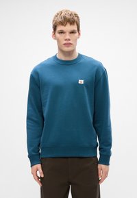 Felpa blu in cotone con collo a giro e polsini a costine. Presenta un piccolo patch con logo sul petto. Texture liscia, vestibilità comoda.