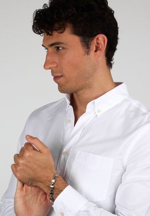 Camicia bianca con bottoni e taschino sul petto, indossata da un uomo, che mostra un bracciale argento con accenti rettangolari al polso.