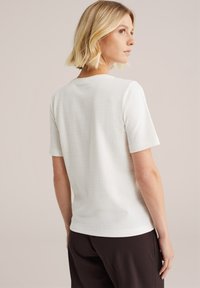 O femeie cu părul blond până la umeri se întoarce, purtând o cămașă albă texturată și pantaloni închiși la culoare, pe un fundal neutru.