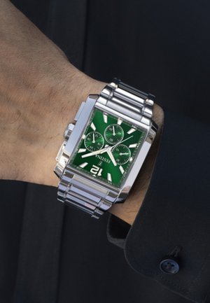 Montre argentée avec un boîtier rectangulaire, un cadran vert, trois sous-cadrans et des index argentés, fixée à un bracelet en maillons métalliques.
