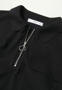 Polo noir en maille avec fermeture éclair argentée et tirette ronde, comprenant une poche poitrine et un col.