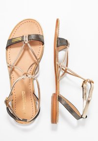 Sandali piatti dorati e neri con cinturini incrociati, soletta in pelle e una leggera suola in legno. Presentano un accento metallico sui cinturini.
