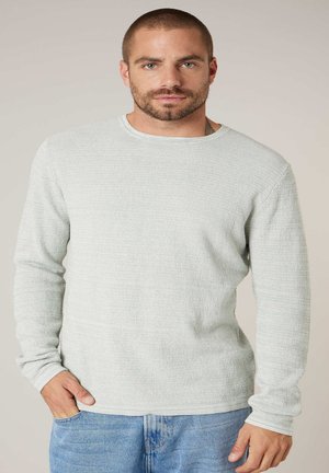 Homme aux cheveux courts et barbe, portant un pull gris clair texturé et un jean bleu, debout avec une main dans la poche.