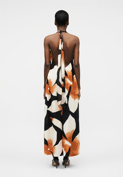 Γυναίκα που φοράει φλοράλ μάξι φόρεμα με λαιμόκοψη halter και ανοιχτή πλάτη, με μεγάλα πορτοκαλί και λευκά λουλούδια, στέκεται μπροστά σε απλό λευκό φόντο.