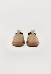 Zapatos de ante beige con acentos oscuros, que cuentan con una tira de agarre y una forma estructurada. La suela es de goma marrón, ofreciendo contraste.