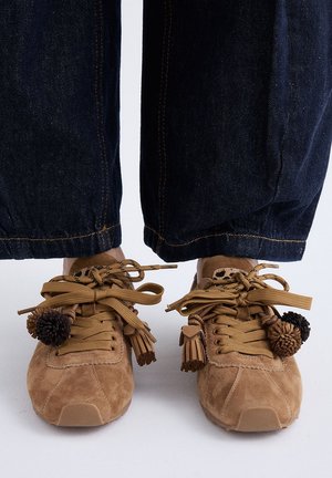 Zapatillas de ante marrón con cordones gruesos color cuero y flecos y pompones decorativos, usadas con vaqueros azul oscuro recortados por encima de los tobillos.