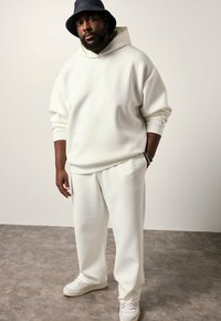 Hvid oversized hoodie med en frontlomme, parret med matchende tætsiddende bukser. Båret med hvide sneakers og en mørk bøttehat. Glat tekstur.