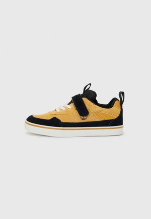 Gul suede-sneakers med svarte detaljer, hvite snøringer og en justerbar stropp. Har en hvit mellomsåle og en pull-tab bak på hælen.