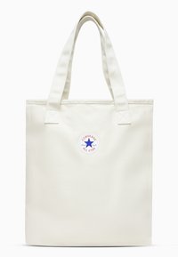 Cream canvas tote bag with two wide handles and a round, colorful Converse All Star logo on the front. Smooth texture, rectangular shape.
