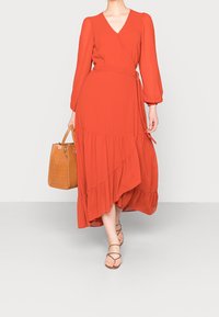 Czerwona sukienka z falbanami, z długimi bufiastymi rękawami, dekoltem w serek, warstwowymi falbanami u dołu, zestawiona z teksturowaną brązową torebką i sandałami na paski.