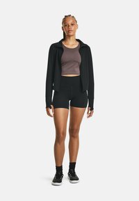 Schwarze Zip-Jacke, eng anliegendes braunes Croptop und hochgeschnittene schwarze Shorts. Das Model trägt schwarze Sneakers mit weißen Sohlen und strukturierten Akzenten.