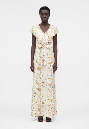 Kvinde iført en ærmeløs, blomsterprikket maxikjole med flæset halsudskæring og bindebånd i taljen, stående mod en ensfarvet hvid baggrund.