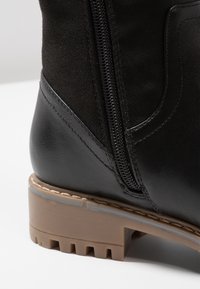 Detailní záběr na černou koženou botu s hnědou podrážkou a bočním zipem na bílé ploše.