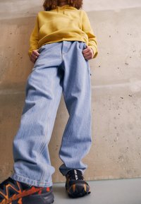 Sudadera con capucha amarilla, vaqueros azul claro de corte relajado y zapatillas negras con detalles en naranja. Fondo de cemento que realza el estilo casual.