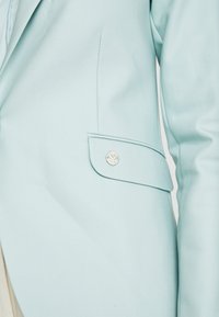 Ljusblå kavaj i slät tyg med skräddarsydd passform, med en knappstängning och en distinkt fickdesign med metalliska detaljer.