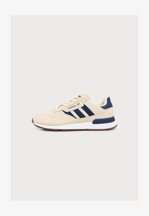 Baskets Adidas beige avec bandes bleu marine, semelle intermédiaire blanche, semelle extérieure marron et détail bleu marine au talon, présentées de profil sur fond blanc.