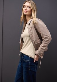 Beige Jacke aus Cord mit Reißverschluss und aufgesetzter Tasche, getragen über einem hellbeigen Oberteil und dunkelblauen Jeans. Glatte Textur, lässiger Schnitt.