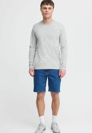 Helles graues Langarmshirt kombiniert mit blauen Denim-Shorts. Das Model trägt weiße Socken und graue Turnschuhe und steht vor einem schlichten Hintergrund.
