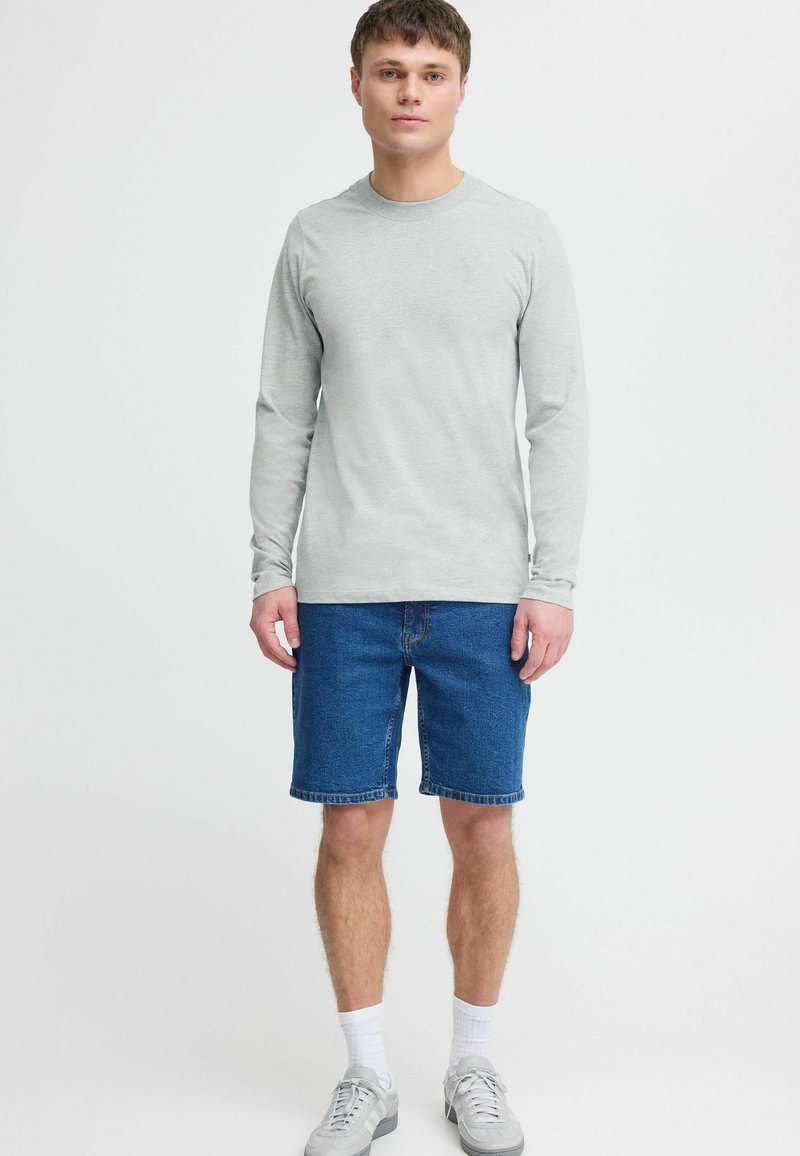 Light grey long-sleeve shirt paired with blue denim shorts. Model wears white socks and grey sneakers, standing against a plain background.