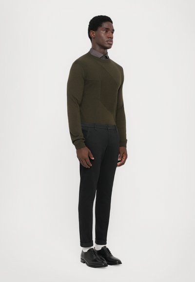 Maglione verde oliva con pannello diagonale, abbinato a pantaloni neri slim e scarpe nere. Presenta una camicia con colletto sotto il maglione.