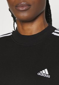 Svart sweatshirt med hög krage, prydd med vita tre-stripsdetaljer på axlarna och en liten vit Adidas-logotyp på bröstet.