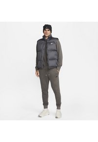 Gilet puffer grigio sopra una felpa scura abbinata a pantaloni jogger. Mostrati i caratteristici loghi Nike. Il modello indossa un berretto nero e sneakers bianche.