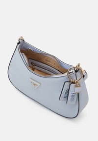 Lyseblå skuldertaske med glat tekstur, guldfarvet hardware, lynlåslukning og en dekorativ mærkat med teksten "GUESS 1981." Indvendigt har den flere rum.