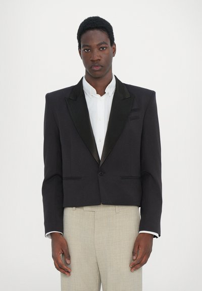 Sort cropped blazer med satin revers, strukturerede skuldre, frontknaplukning og to forlommer, båret med en hvid skjorte.