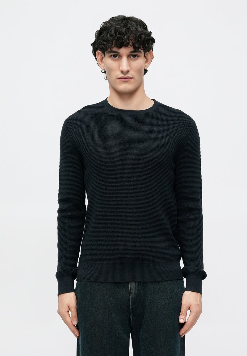 Suéter de punto azul marino con un patrón texturizado, cuello redondo y mangas largas. Combinado con pantalones de mezclilla oscuros para un look casual.