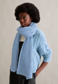 Jasnoniebieski dziergany szalik z miękkiego materiału, zestawiony z dopasowanym swetrem o podobnym kolorze i teksturze, prezentujący prążkowane detale.