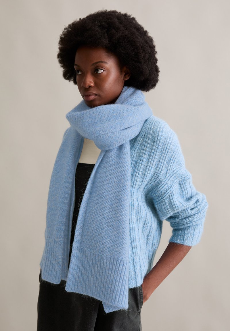 Jasnoniebieski dziergany szalik z miękkiego materiału, zestawiony z dopasowanym swetrem o podobnym kolorze i teksturze, prezentujący prążkowane detale.