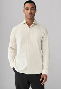 Chemise beige clair à manches longues avec un devant à boutons, col classique et texture douce. Associée à un pantalon noir.