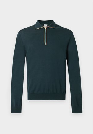 Homme portant un haut sombre à manches longues avec col zippé, un pantalon noir à jambes larges et des chaussures style mocassin marron clair, debout sur fond blanc.