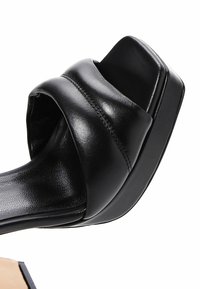 Sandale en cuir noir à talon block, avec une large bride supérieure, texture lisse et design à bout carré. La semelle est élégante et légèrement surélevée.