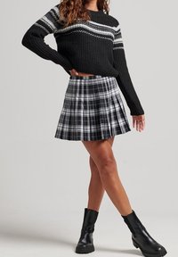 Schwarzer Strickpullover mit grauen Streifen an den Schultern, kombiniert mit einem schwarz-weißen karierten Plisseerock. Das Modell trägt schwarze Stiefeletten.