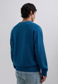Homme avec des cheveux courts et bouclés portant un sweat bleu et un jean bleu clair, de dos contre un fond clair.