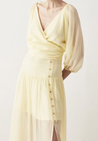 Keltainen mekko, jossa on kietaisumallinen yläosa, volyymikkäät hihat, kerroksellinen helma ja koristeelliset kultanapit sivusaumassa.