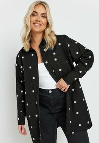 Camicia nera con bottoni, caratterizzata da ricami bianchi a forma di margherita, maniche lunghe e una vestibilità comoda. Indossata sopra un top bianco e pantaloni neri.